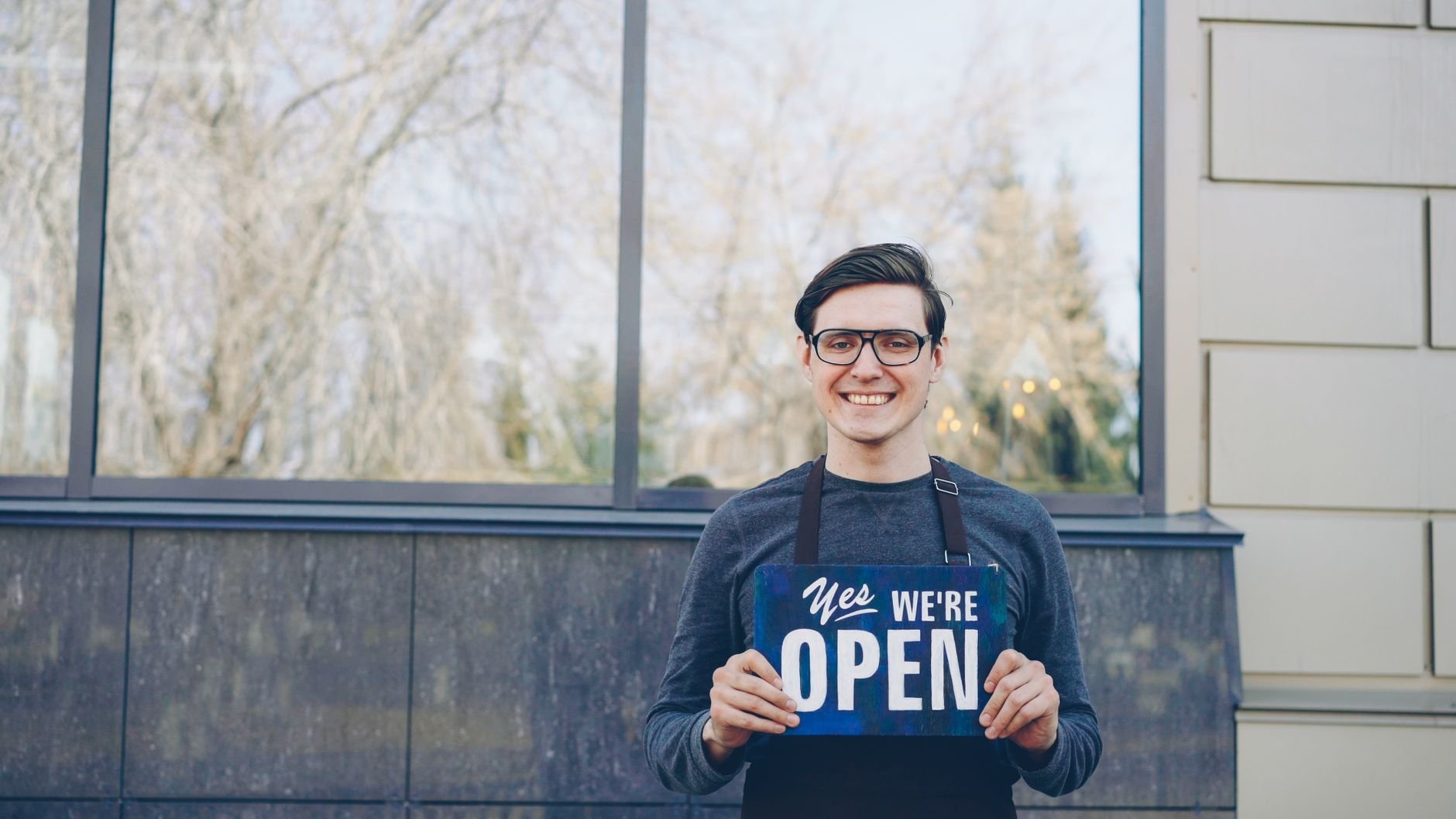 Smiling young business owner in glasses holding a 'Yes, we're open' sign in front of their store